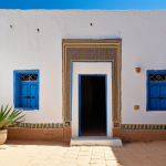**Prompt:** Traditional Libyan house, white walls, central courtyard with plants, small windows, bright sunlight, clear blue sky, desert background, sense of coolness and tranquility.